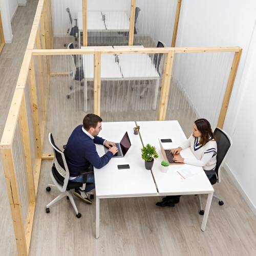 Dos personas trabajando en laptops sentadas en escritorios separados por divisores de madera y plástico dentro de un espacio de coworking.