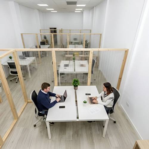 Dos personas trabajando con laptops en escritorios separados por divisores de madera y vidrio dentro de un espacio de coworking moderno.
